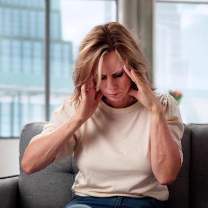 A woman sitting on a couch, pressing her temples as if suffering from a headache.