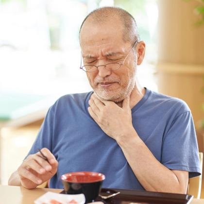 Elderly man with glasses looking concerned while touching his throat and holding a spoon over a meal in a bright dining area.