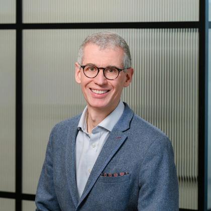 Professional portrait of a smiling man with glasses wearing a blue blazer and light blue shirt, standing in front of a patterned glass backdrop.
