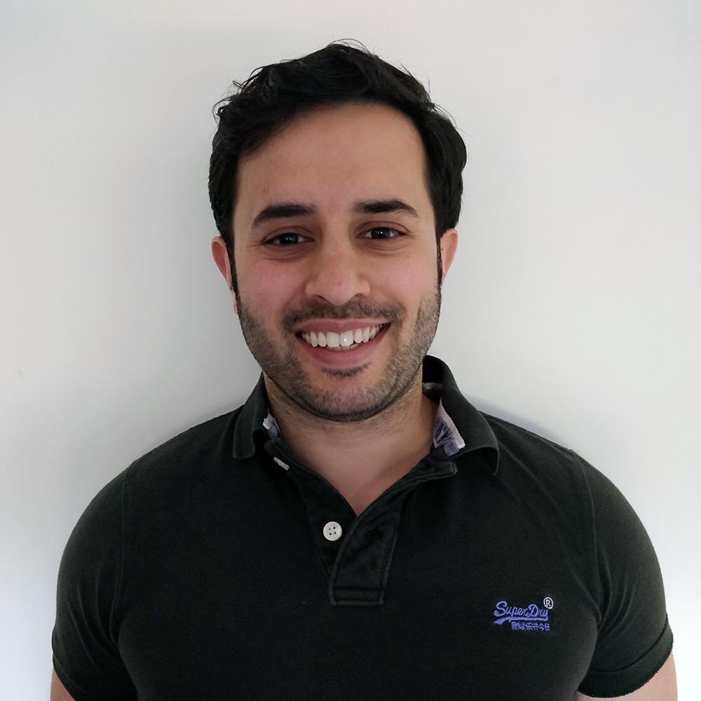 Smiling man in a black polo shirt standing against a white wall.