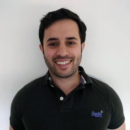 Smiling man in a black polo shirt standing against a white wall.