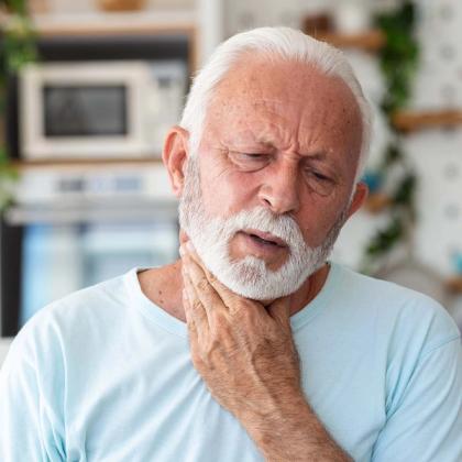 Elderly man with white beard experiencing throat pain or discomfort in his kitchen