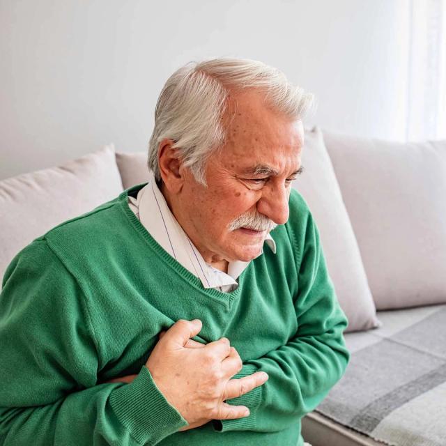 An elderly man in a green sweater clutches his chest with a pained expression.