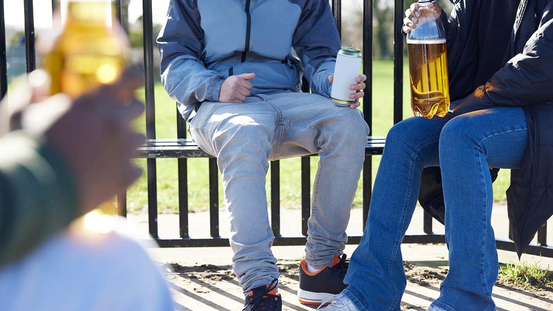 Three people sit on an outdoor bench holding cans and bottles of alcoholic drinks.