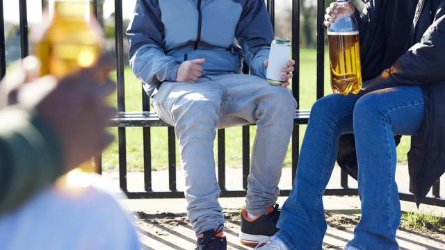 Three people sit on an outdoor bench holding cans and bottles of alcoholic drinks.