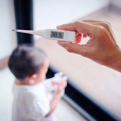 Digital thermometer displaying a fever temperature of 38.9°C held by an adult hand with a toddler in the background