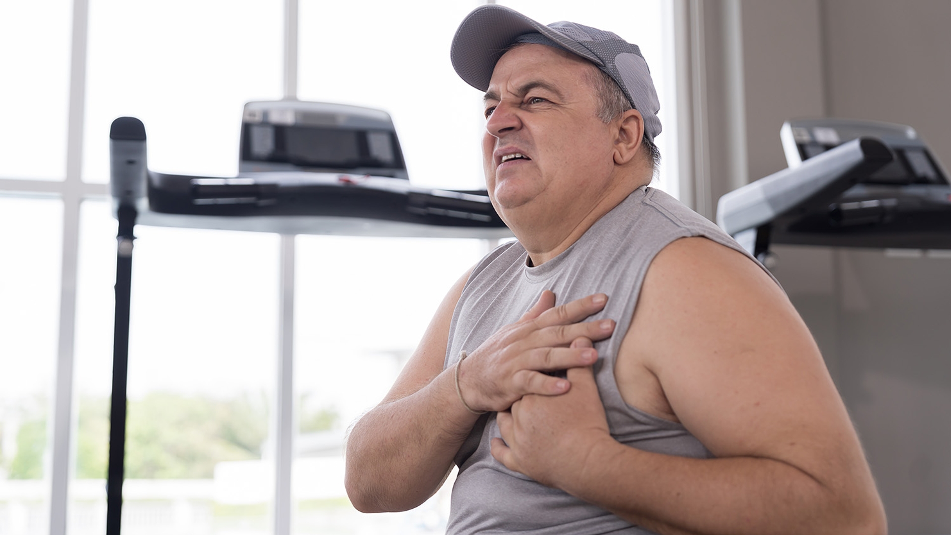 Overweight man in a gym clutches his chest, grimacing in pain.