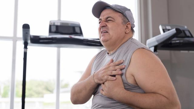 Overweight man in a gym clutches his chest, grimacing in pain.