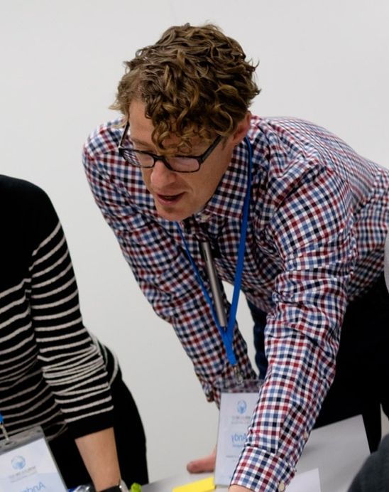 Picture of Andy leaning over a table consulting with others on a piece of work. 