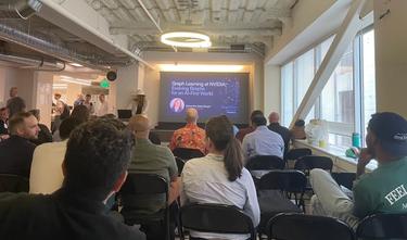 A view of a presenter and her title slide from six rows back in an office space turned small conference venue. 