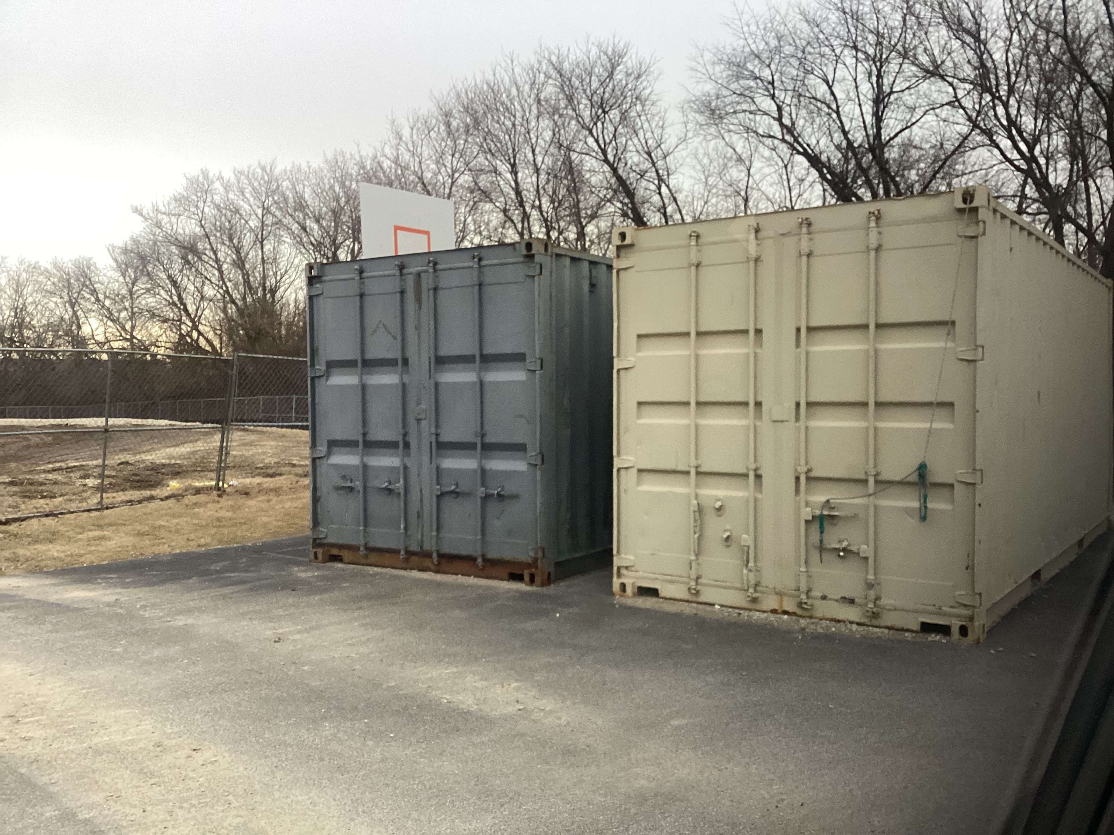 Two shipping containers on a paved site with closed end doors and locking bars visible