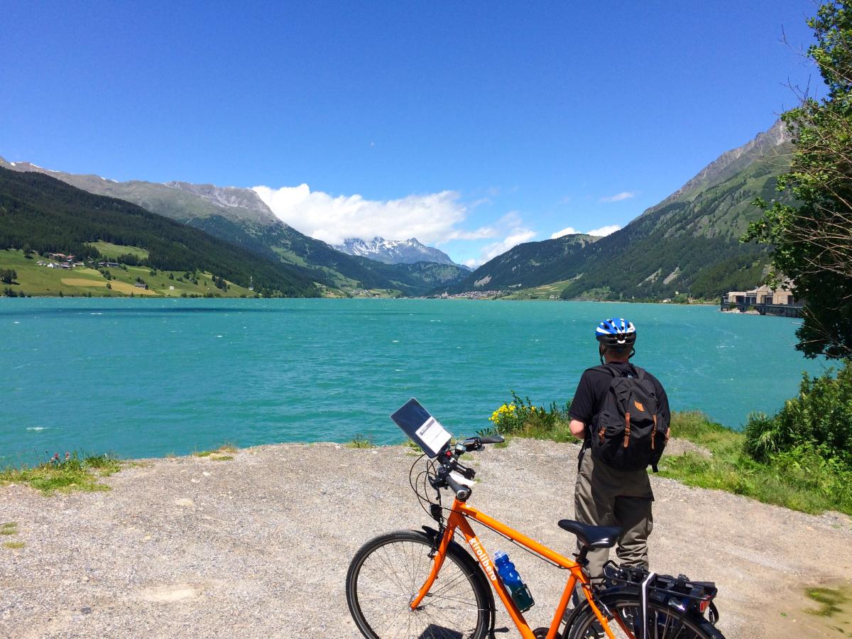 a cyclist on the Adige River Cycle Route (also known as Etschradweg) in Italy, likely near the Reschensee (Lake Reschen) in South Tyrol