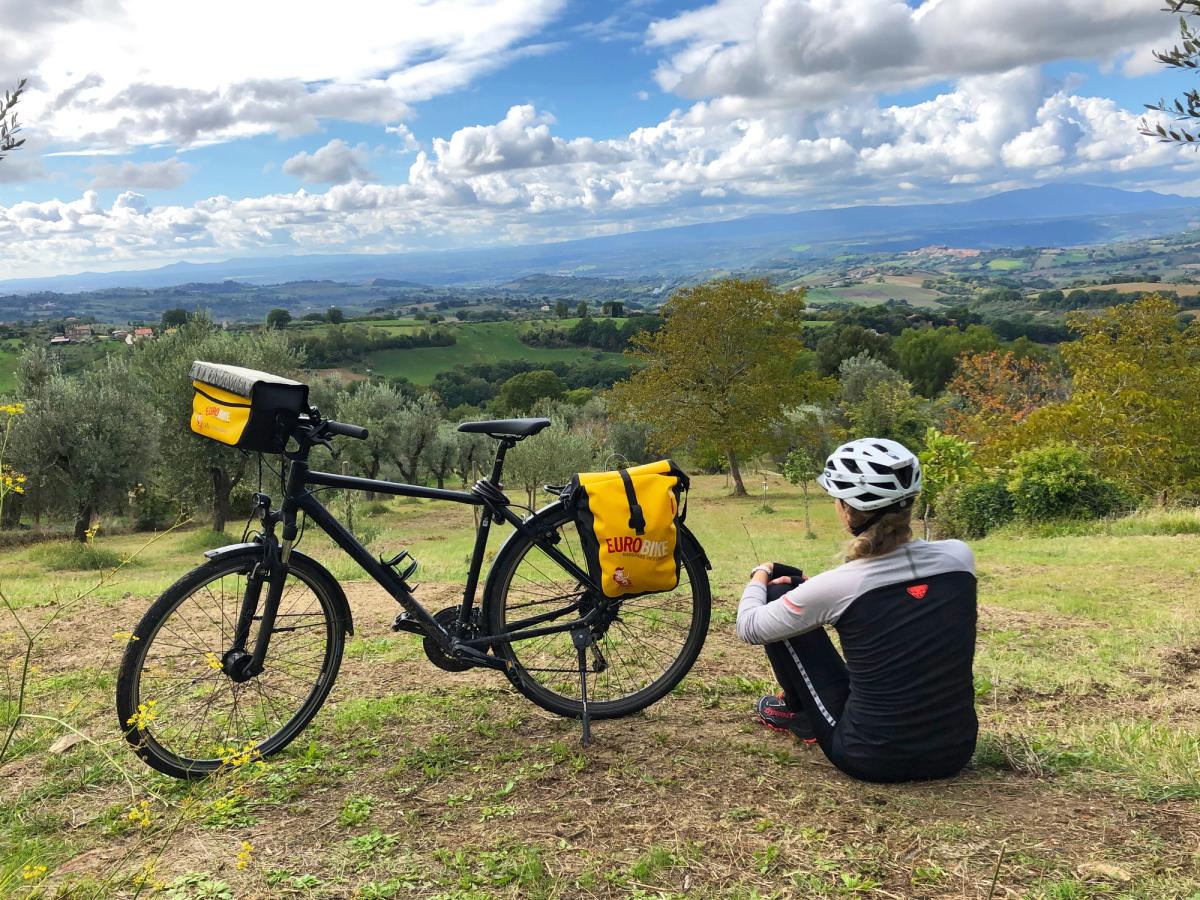 a cyclist taking a break during a bike tour, likely in a scenic, hilly region with olive groves or vineyards, possibly in Italy