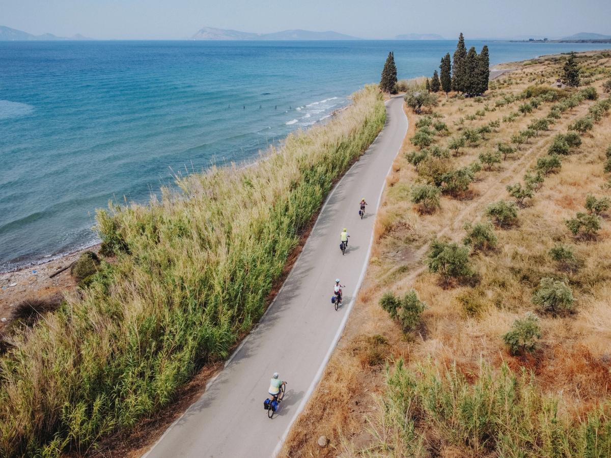 a group of cyclists on a coastal road, likely in the Saronic Gulf region of Greece, possibly as part of a cycling tour like the Laconia Coastal Biking Tour