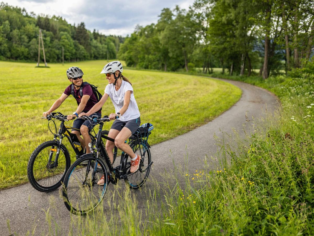 a man and a woman riding bicycles on a paved path through a rural, grassy landscape