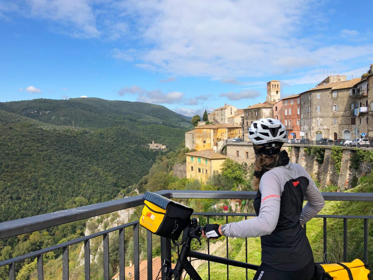 a cyclist on a bike tour, likely in the Umbria region of Italy, overlooking a picturesque hill town like Narni or a similar village in Tuscia