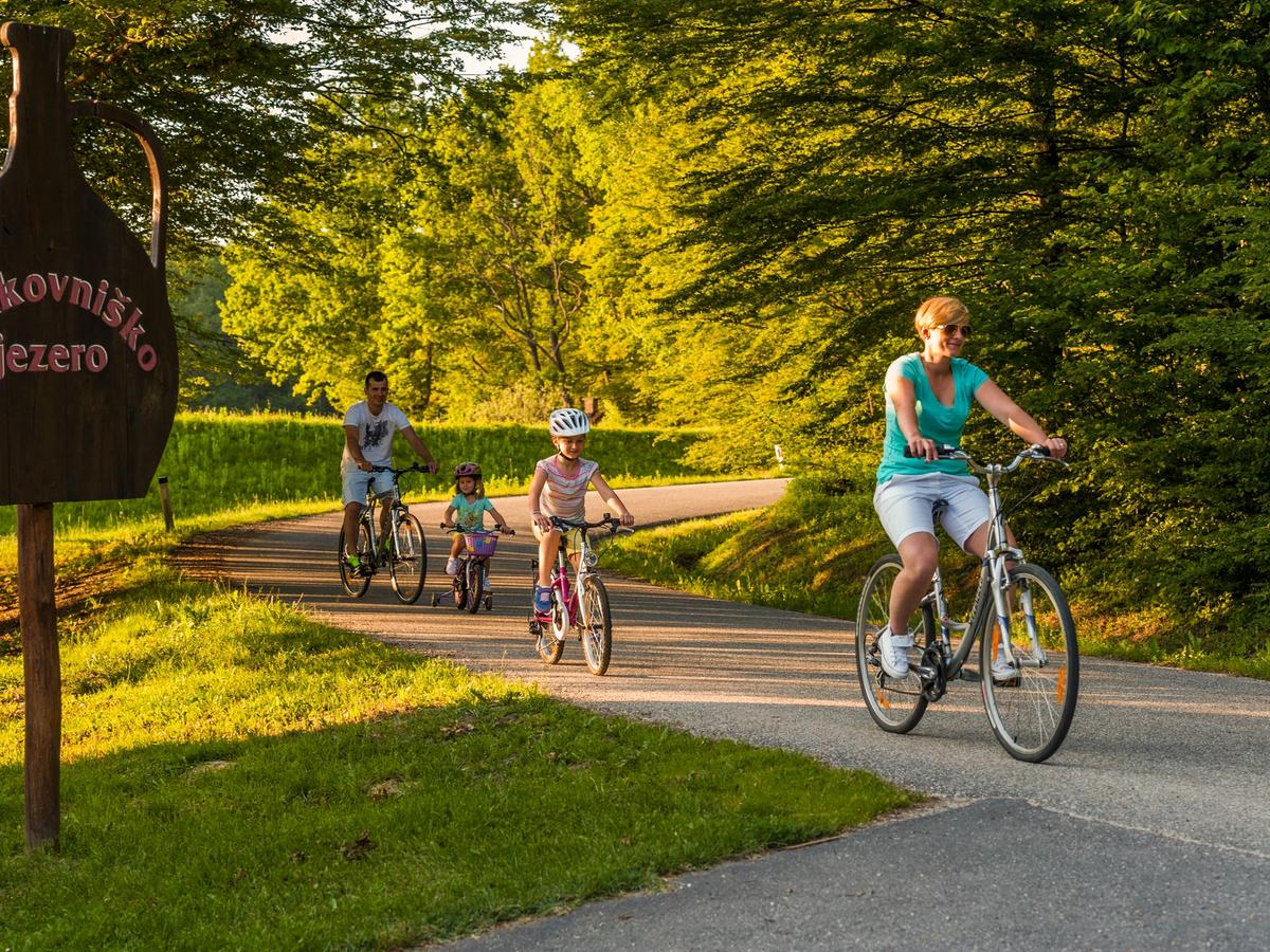 a family cycling on a path near Bukovniško jezero (Bukovnica Lake) in Slovenia. The area is part of Terme Lendava, a thermal resort known for its spa and outdoor activities