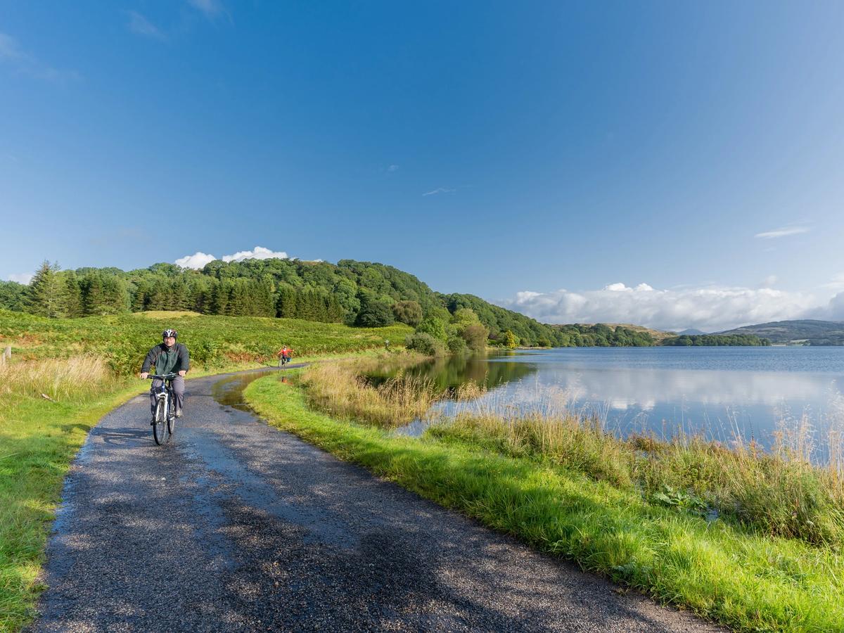 a person cycling along a scenic path next to a body of water, likely a loch or lake, in a natural setting