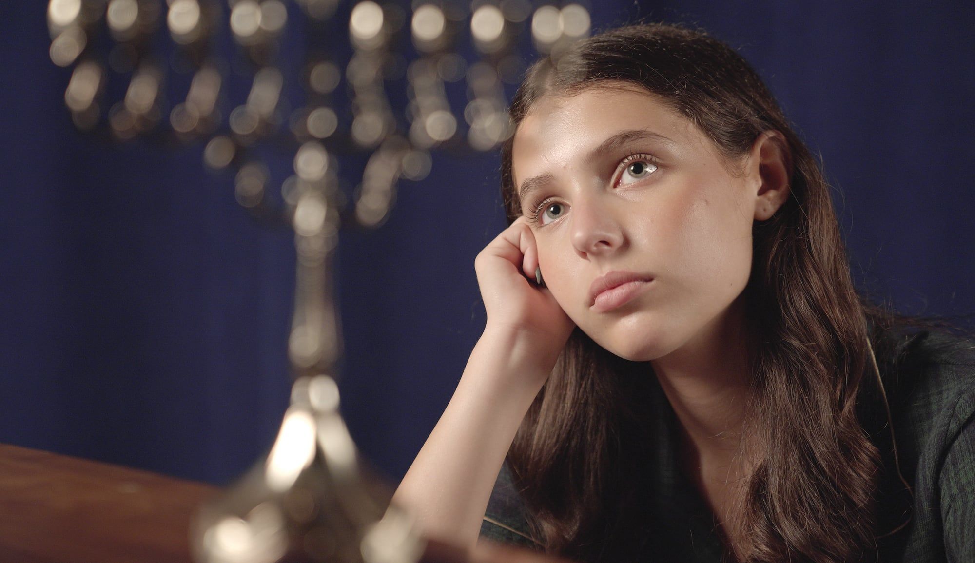Sasha Bloom as Anne Frank, seated beside a menorah