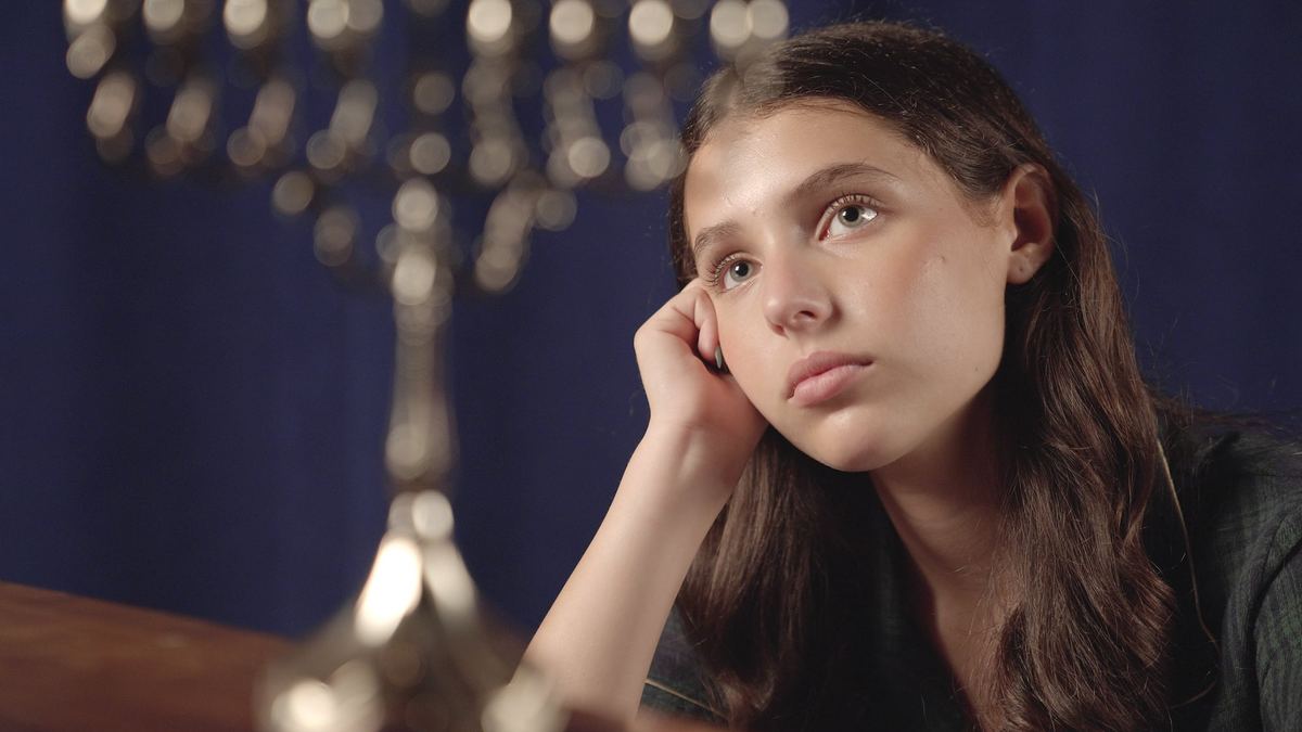 Sasha Bloom as Anne Frank, seated beside a menorah