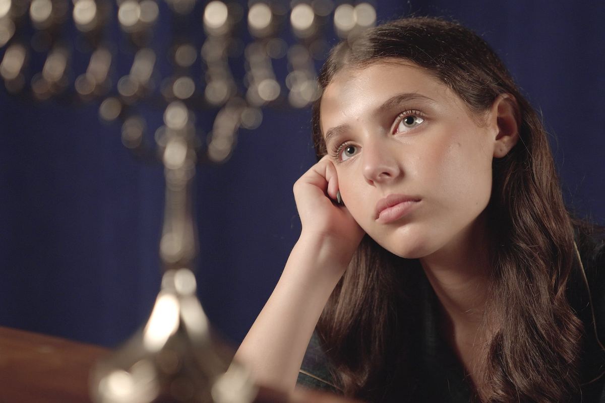 Sasha Bloom as Anne Frank, seated beside a menorah