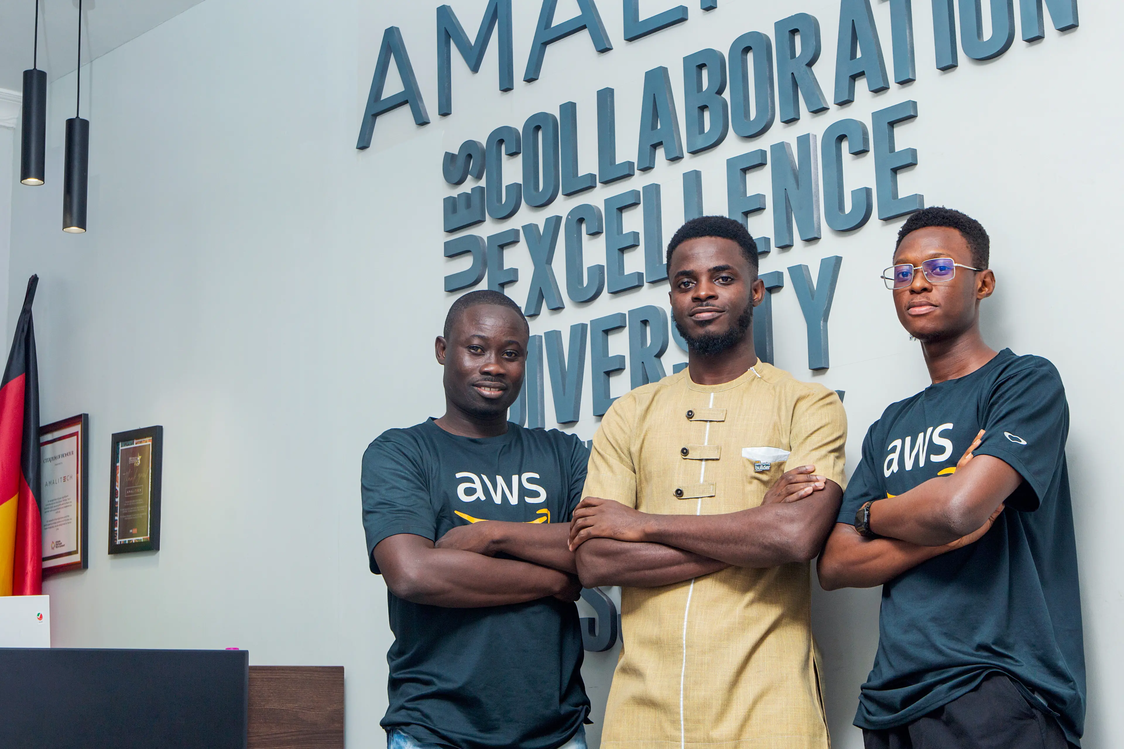 Three men standing in front of the AmaliTech logo which is placed on the wall, crossing their arms. Two are wearing AWS shirts