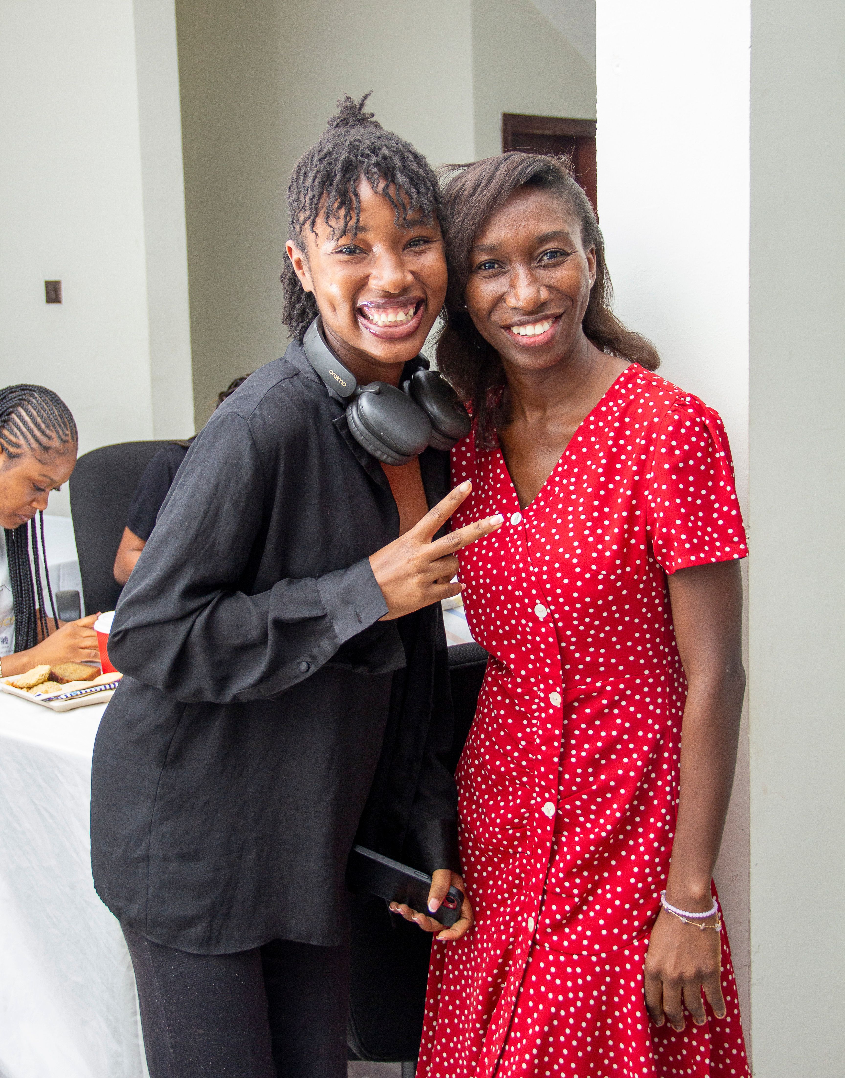 Two women smiling at the camera. The women on the left side is making a peace sign with her hand and wearing headphones around her neck.