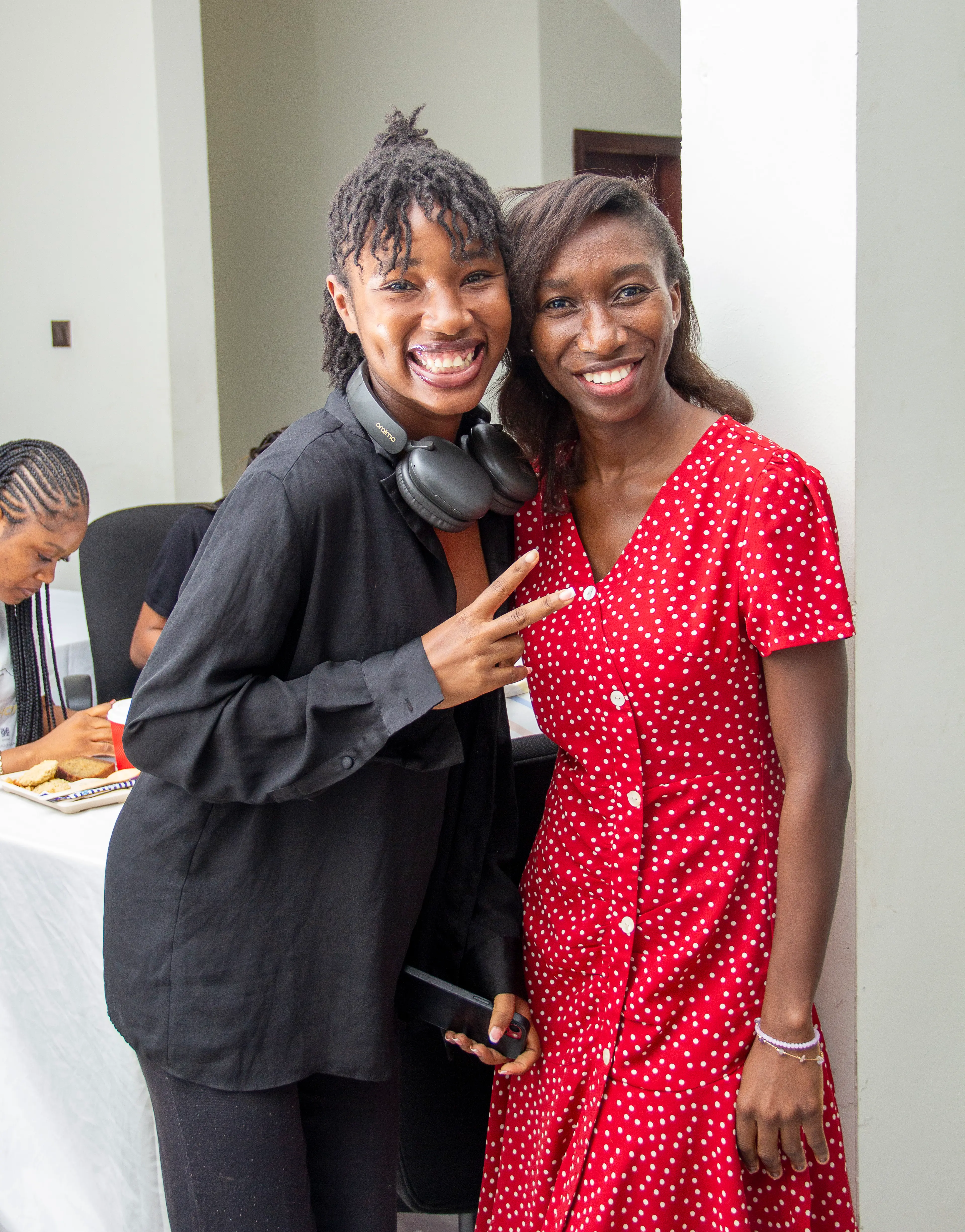 Two women smiling at the camera. The women on the left side is making a peace sign with her hand and wearing headphones around her neck.
