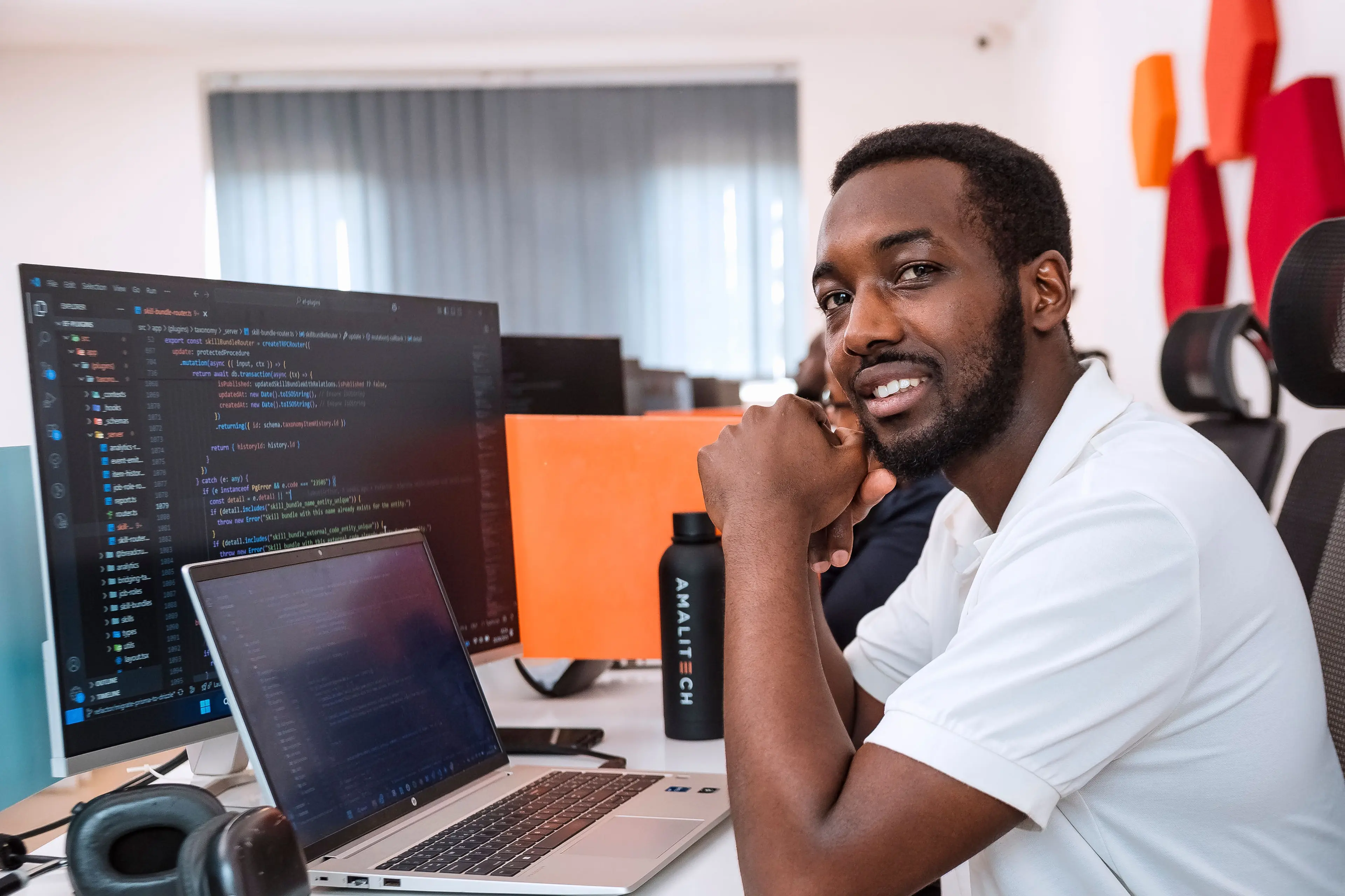 A man sitting at his desk which is filled with a laptop, monitor and headphones, smiling at the camera