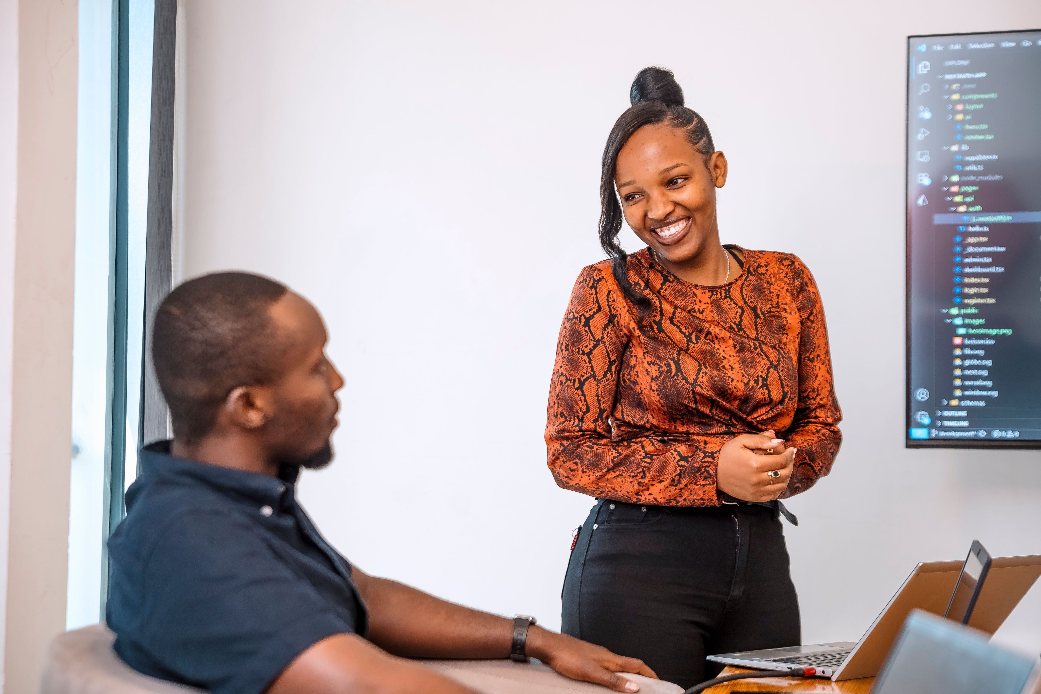A woman standing and presenting while smiling at her colleague who is sitting in front of her
