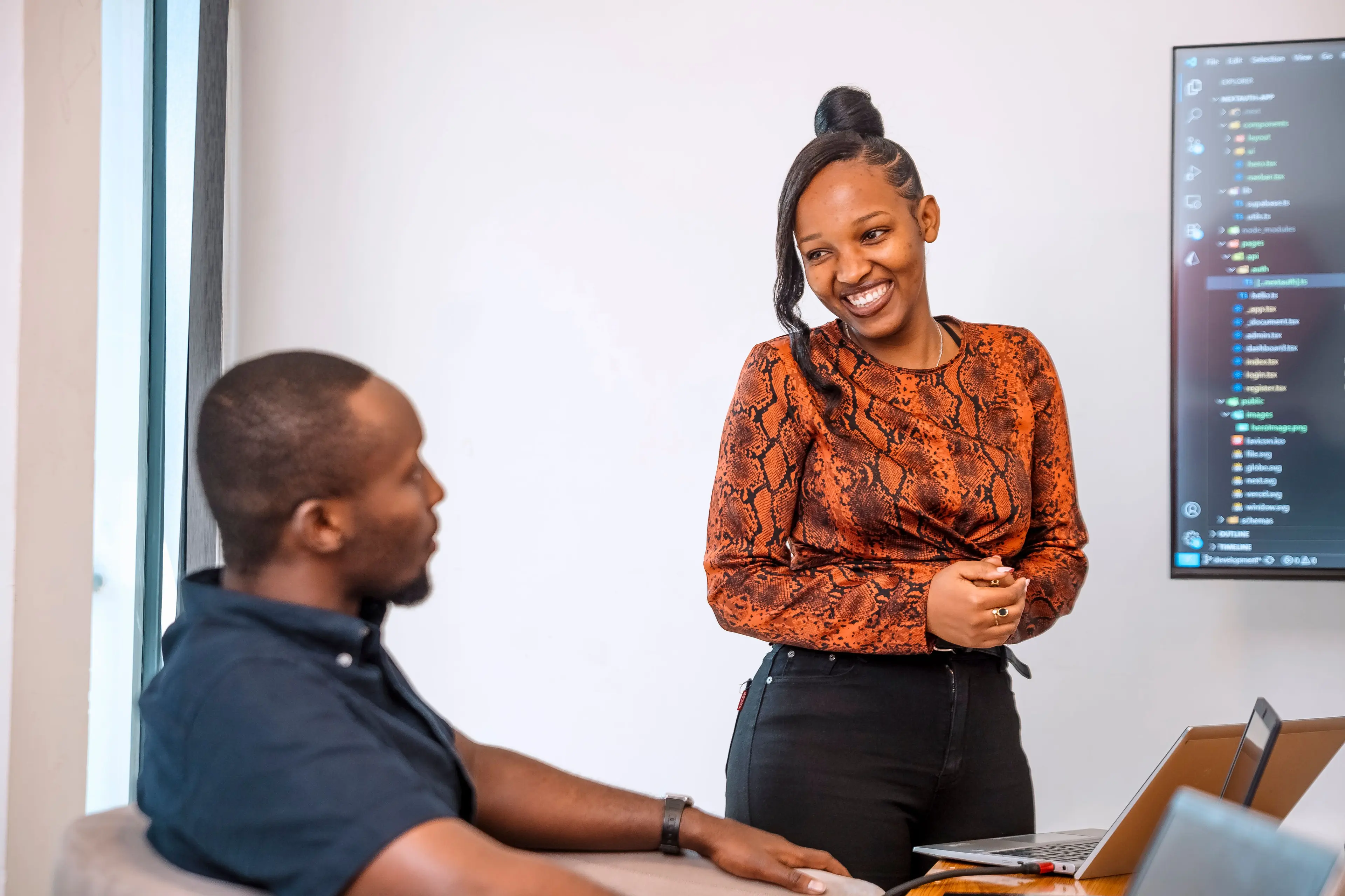 A woman standing and presenting while smiling at her colleague who is sitting in front of her