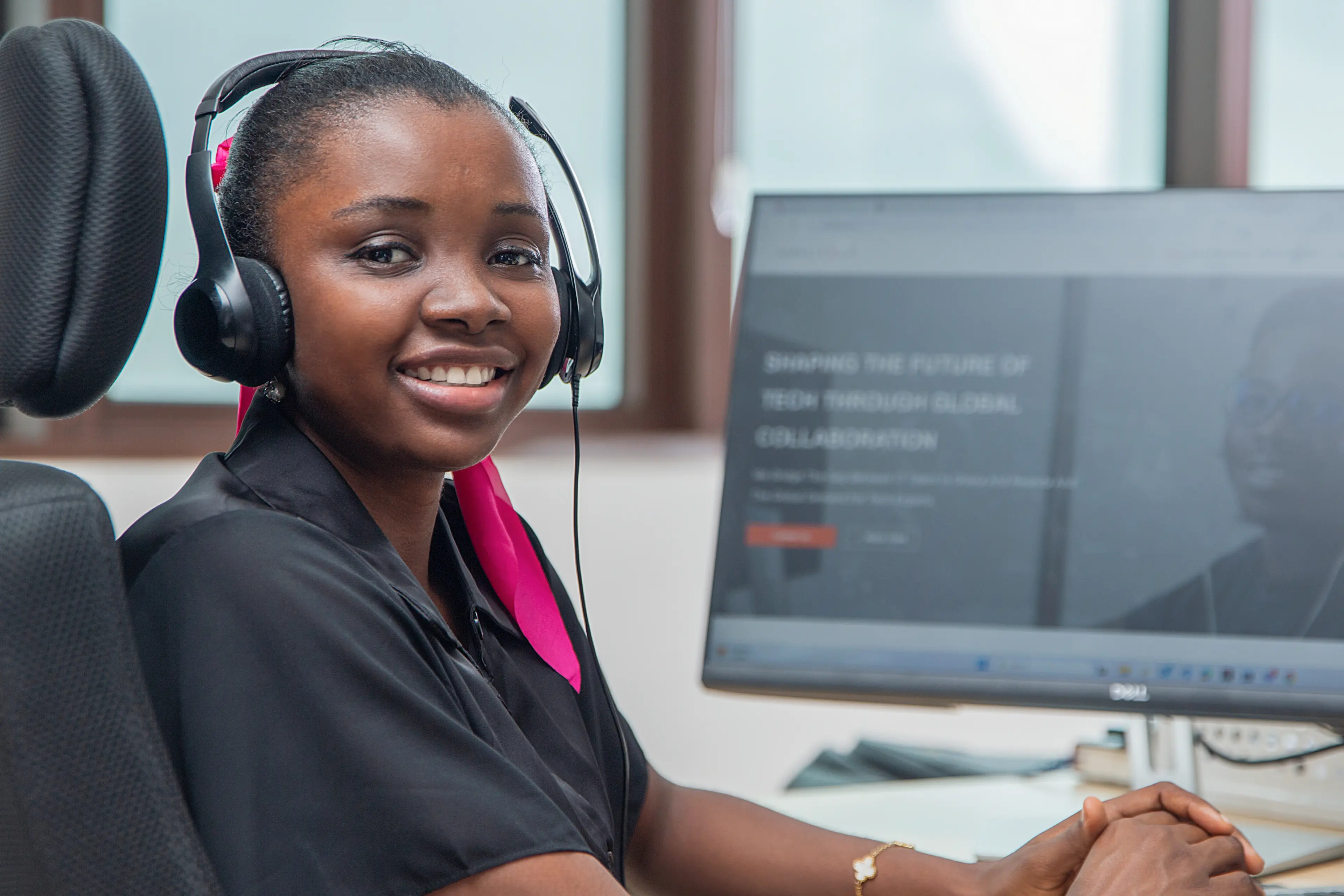 A women smiling at the camera, sitting at her desk. You can see her computer on the desk.