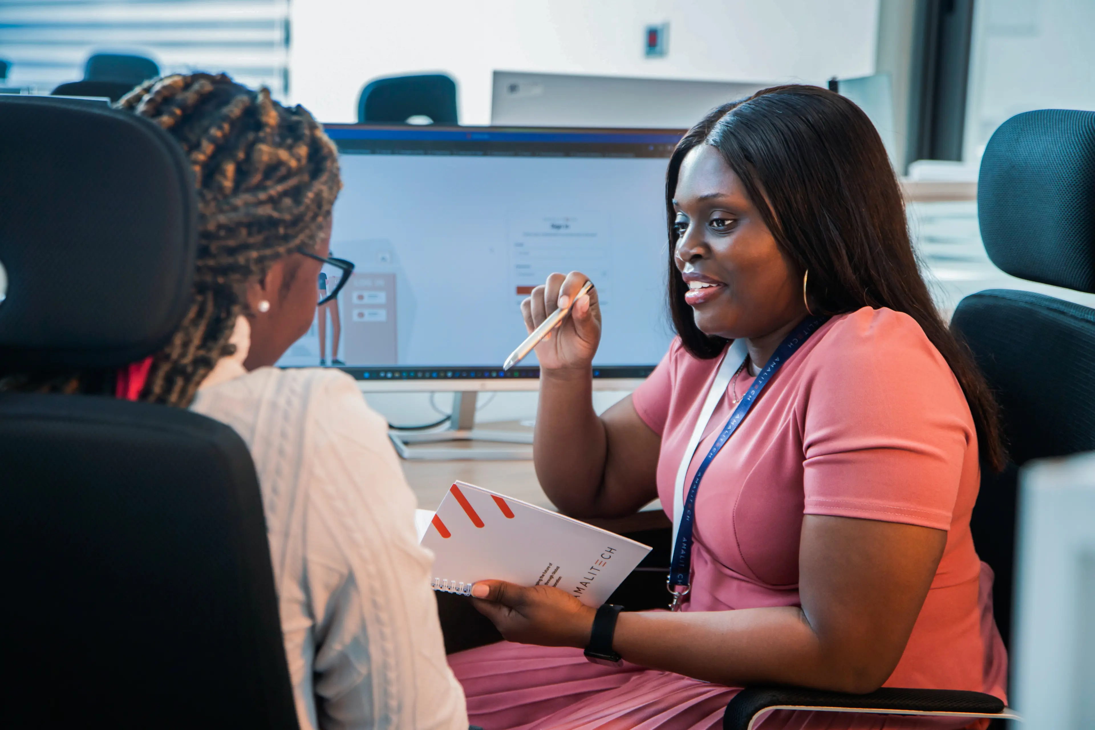 two women speaking at a desk. One is holding a notepad and pen