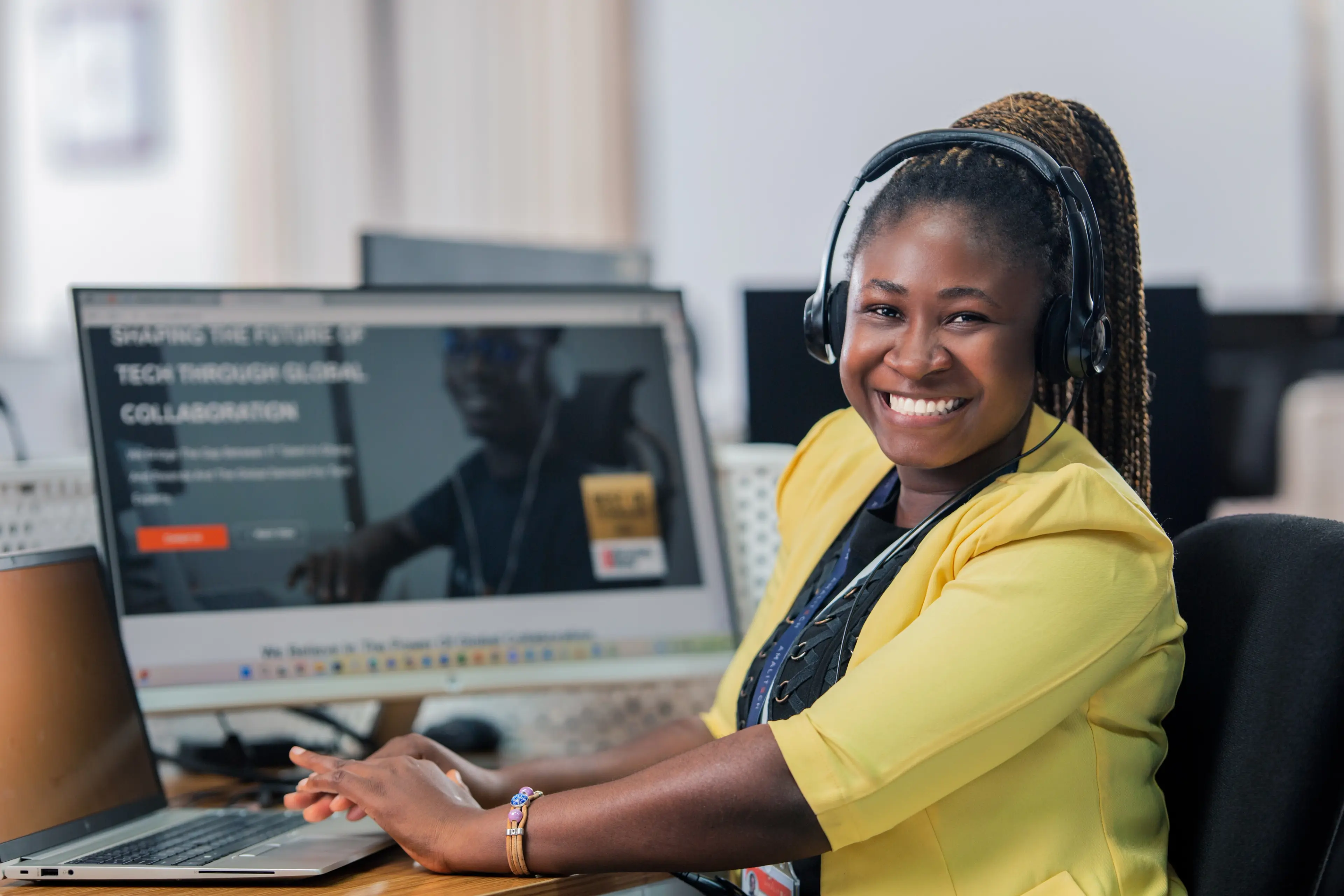 A women wearing a yellow blazer, sitting at her desk in front of a computer and smiling at the camera