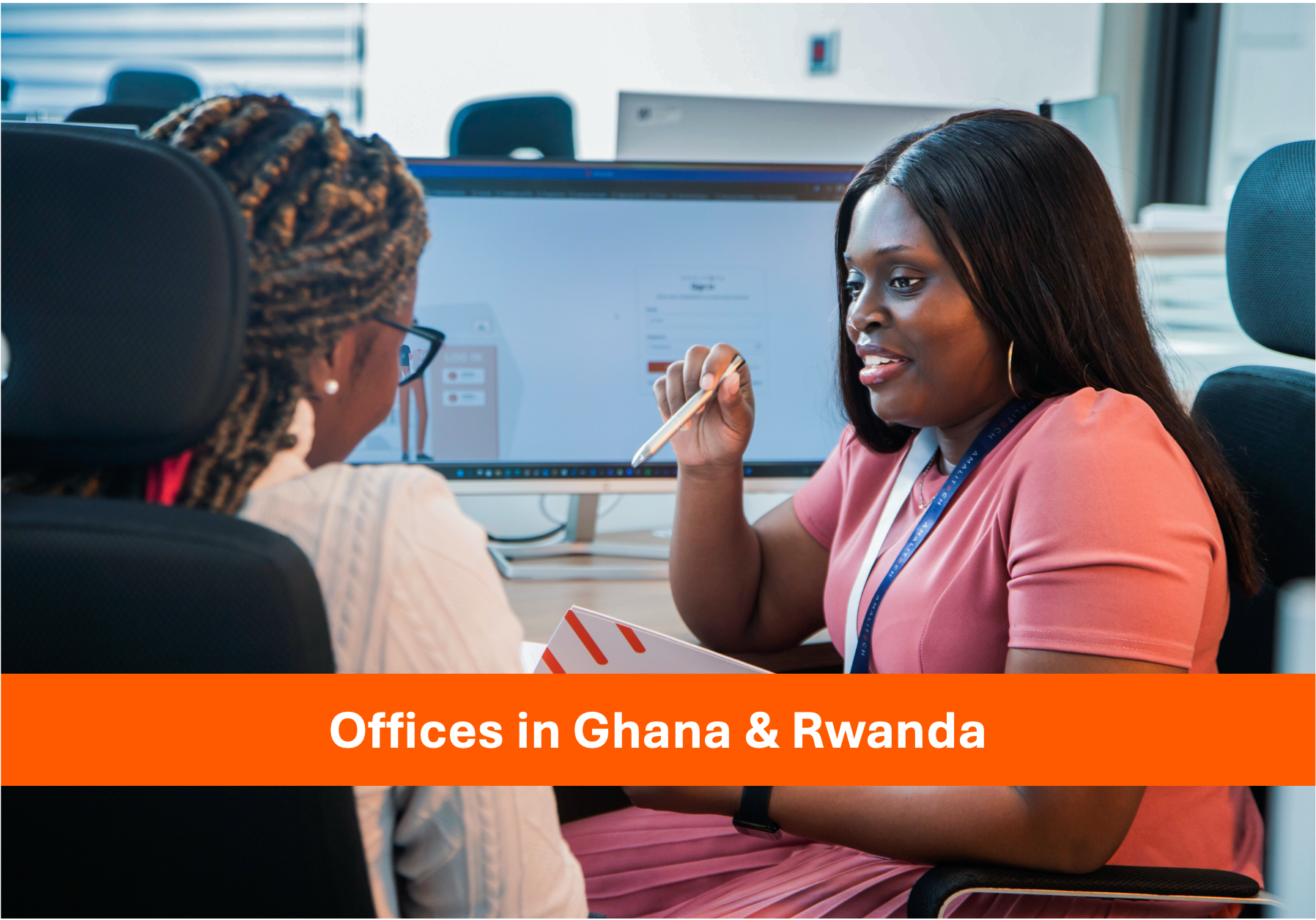 Two women sitting in front of a computer, talking. One women is holding a pen and notepad in her hands.