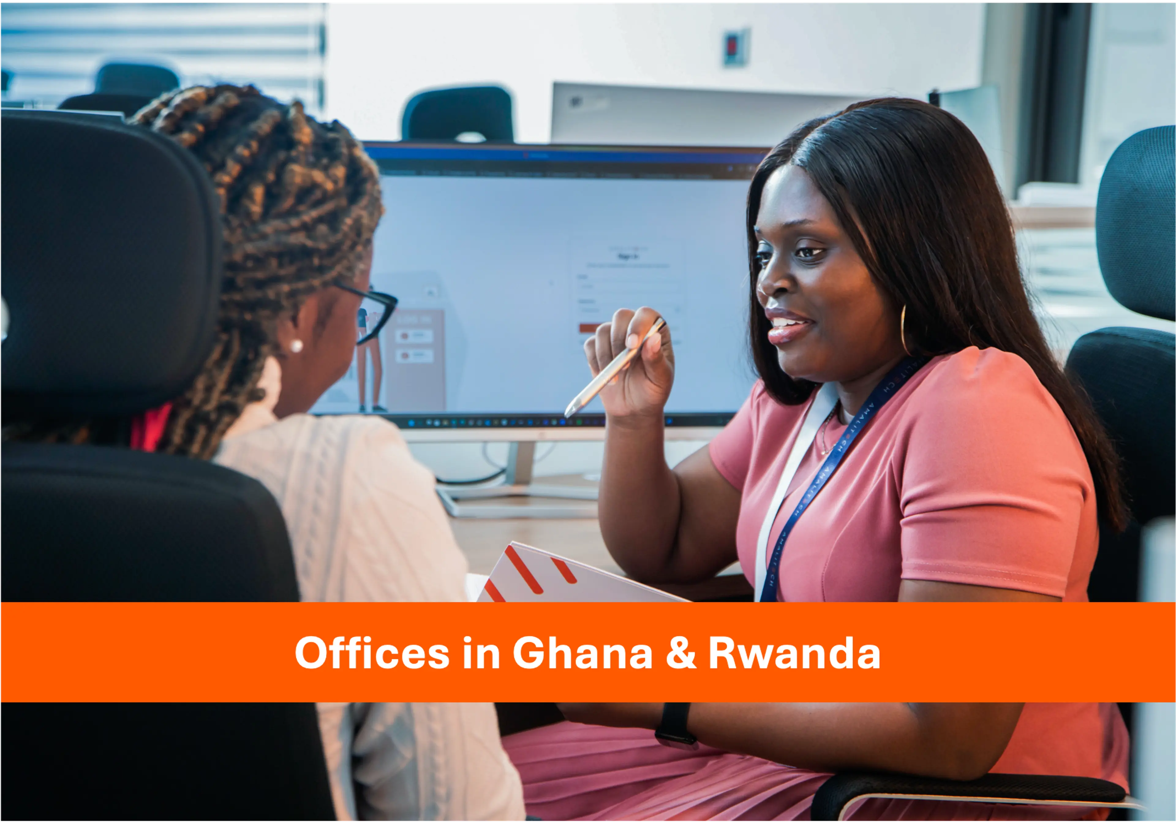 Two women sitting in front of a computer, talking. One women is holding a pen and notepad in her hands.
