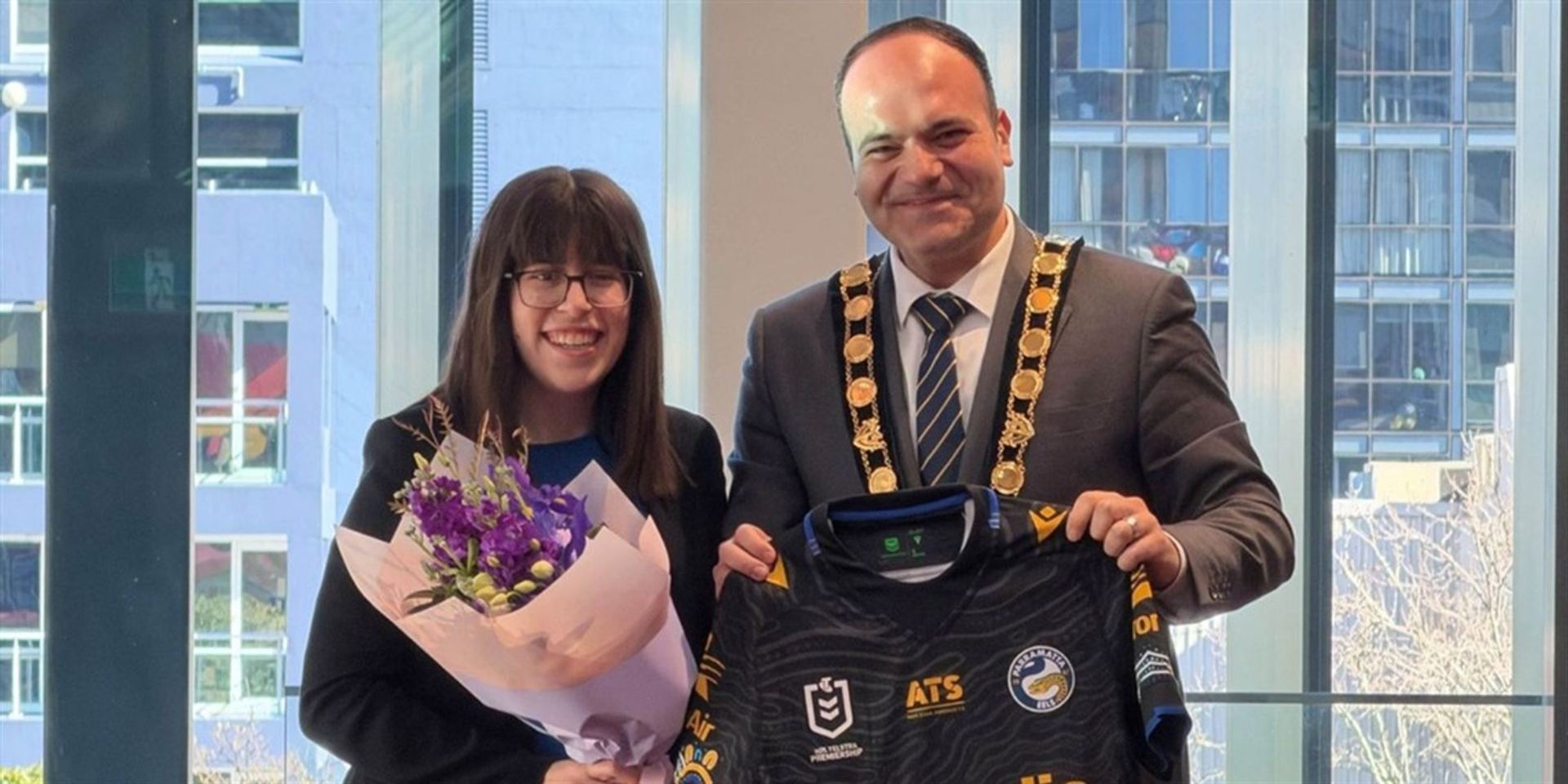 Yasmine smiling with a bouquet of purple flowers beside a man in a ceremonial chain of office who holds up a Parramatta Eels jersey