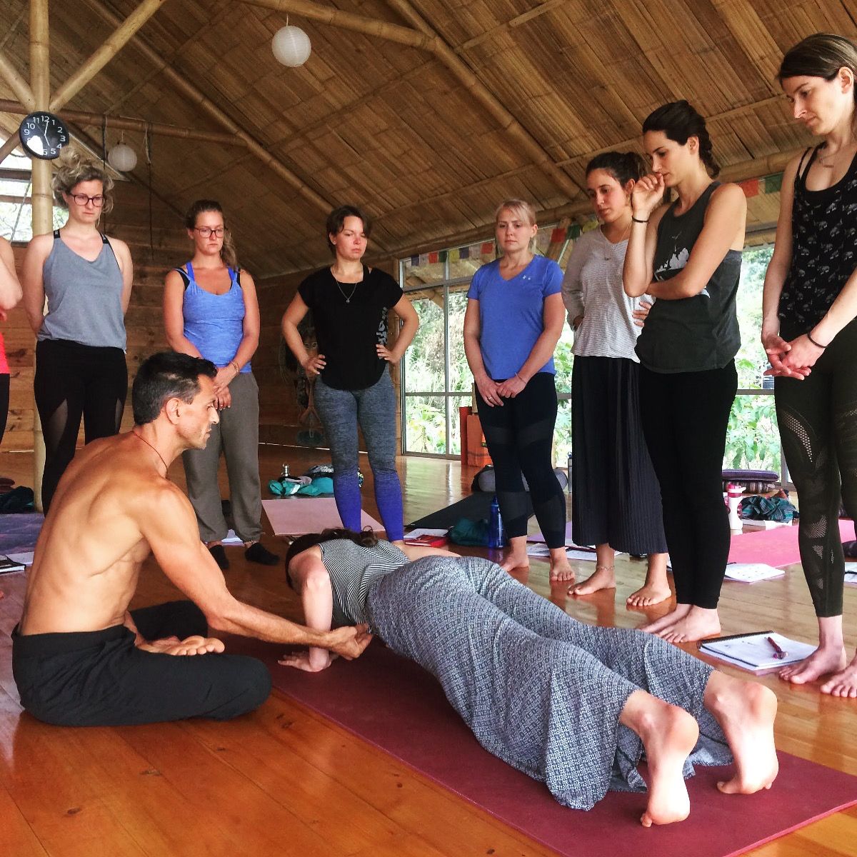 A teacher kneeling at the center while a circle of trainees watches him adjust a student.
