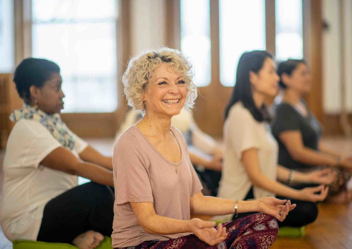 An older woman smiling, seated in a yoga class.