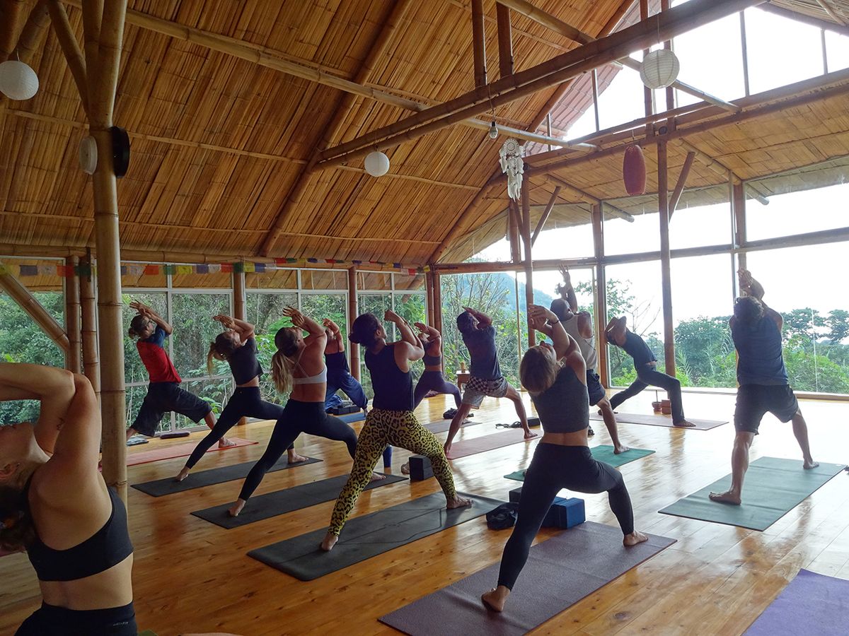 A class holding warrior pose in a bright wooden shala.