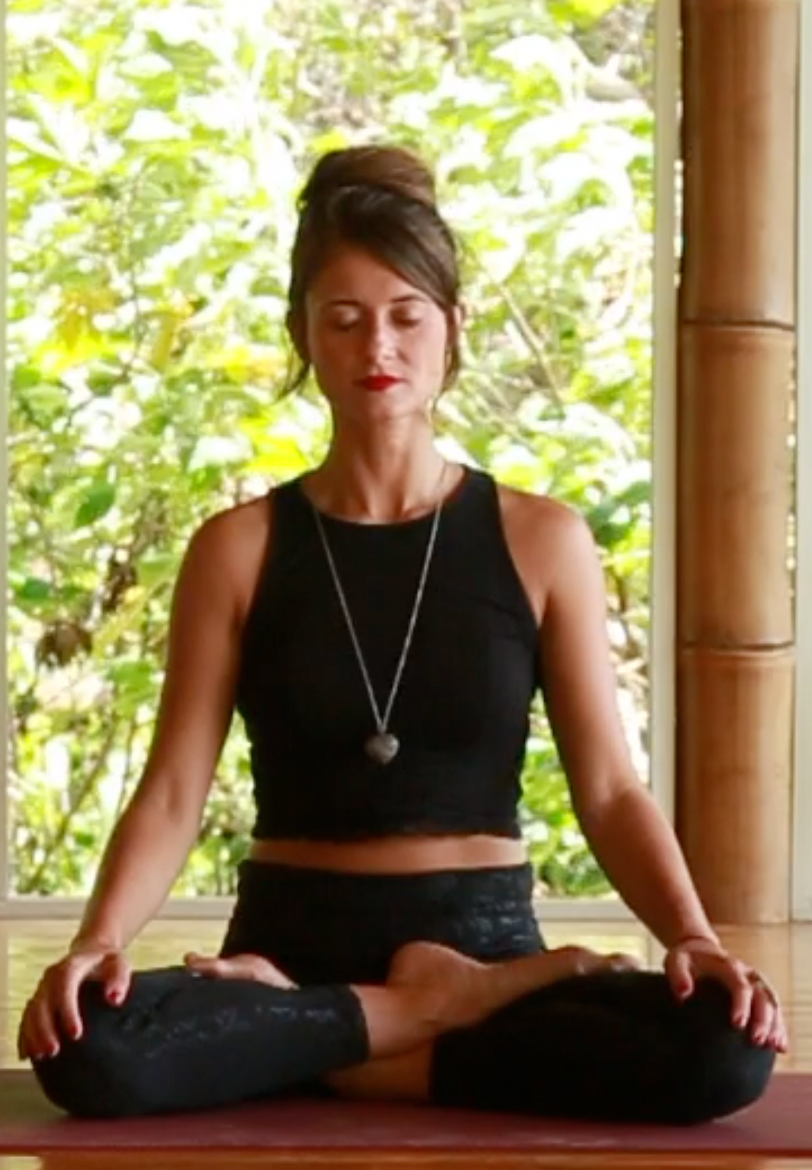 A woman seated with her eyes closed, hands resting on her knees.