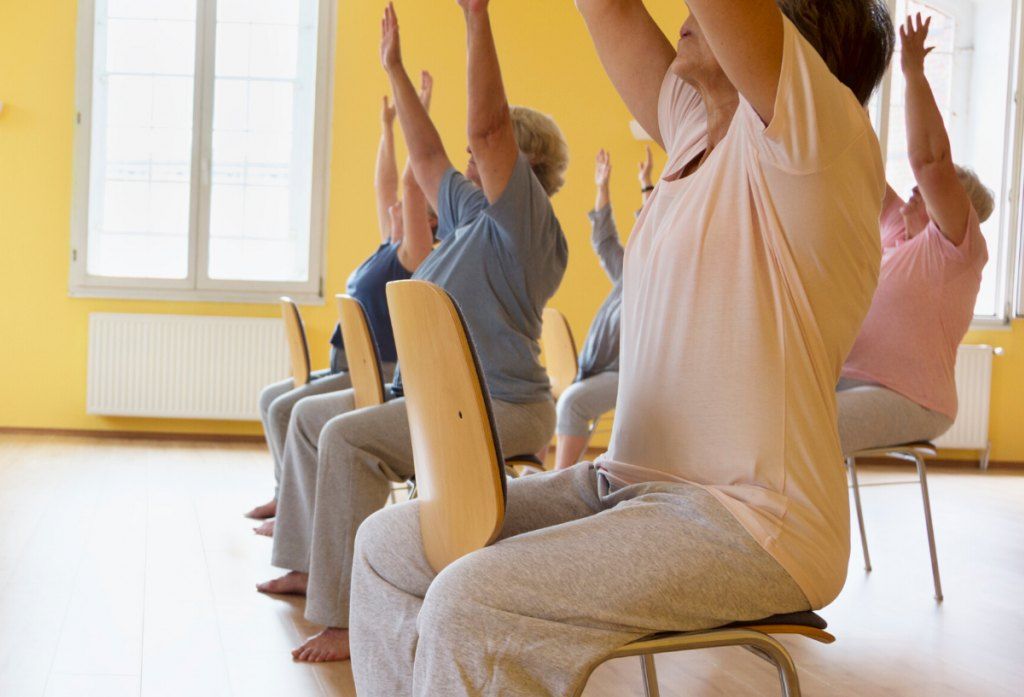 Older students seated on chairs, arms lifted in a sun salutation.