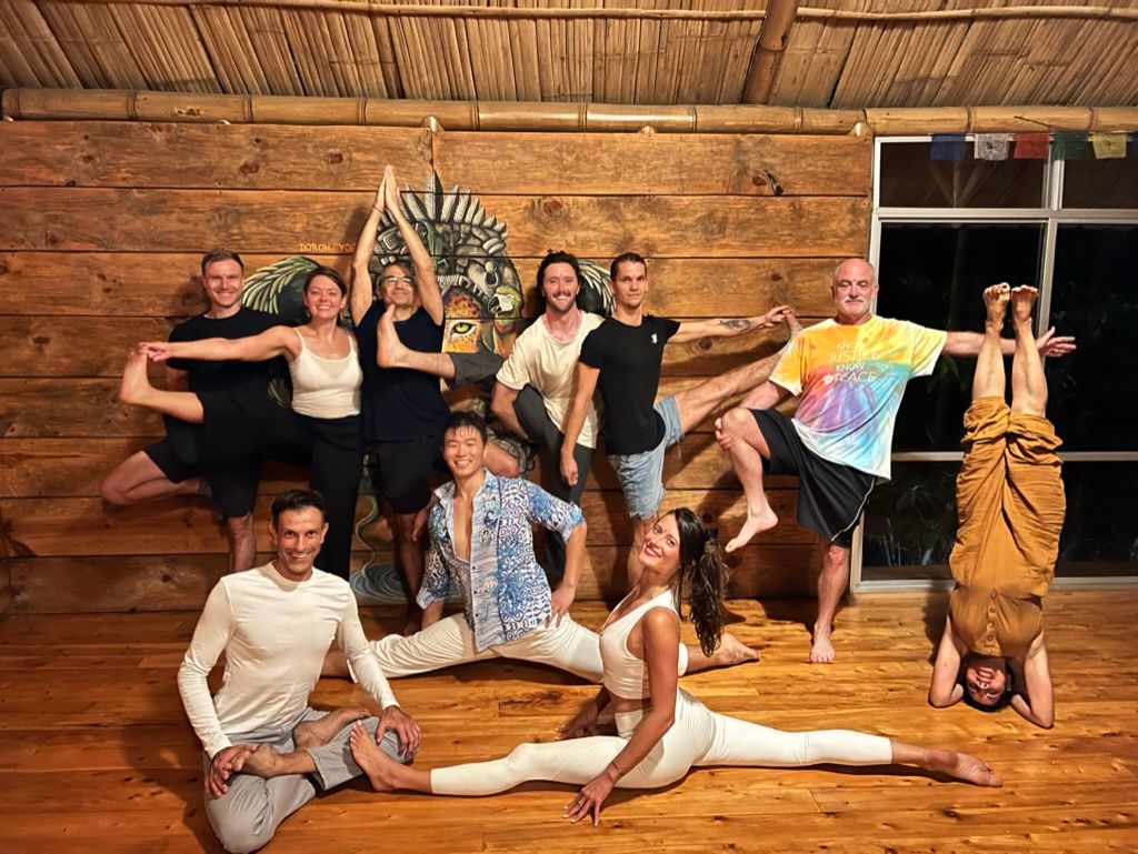 A group laughing together in a wooden yoga room, some in tree pose and a split.