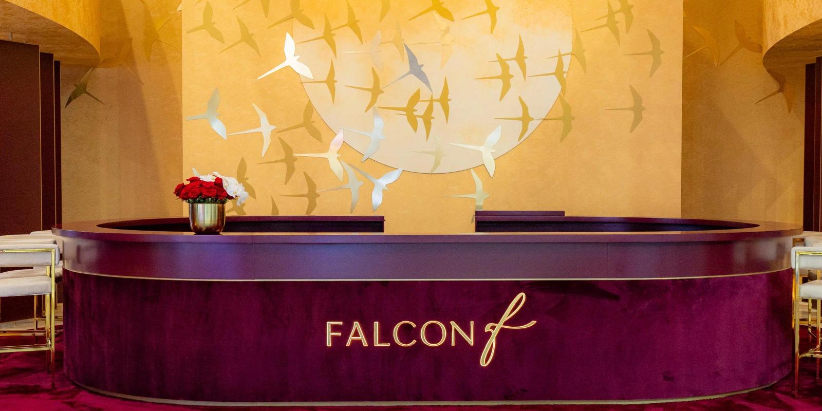 Burgundy velvet reception desk with backlit Falcon lettering and a vase of red roses, in front of a gold wall of metal bird silhouettes on a Falcon exhibition stand