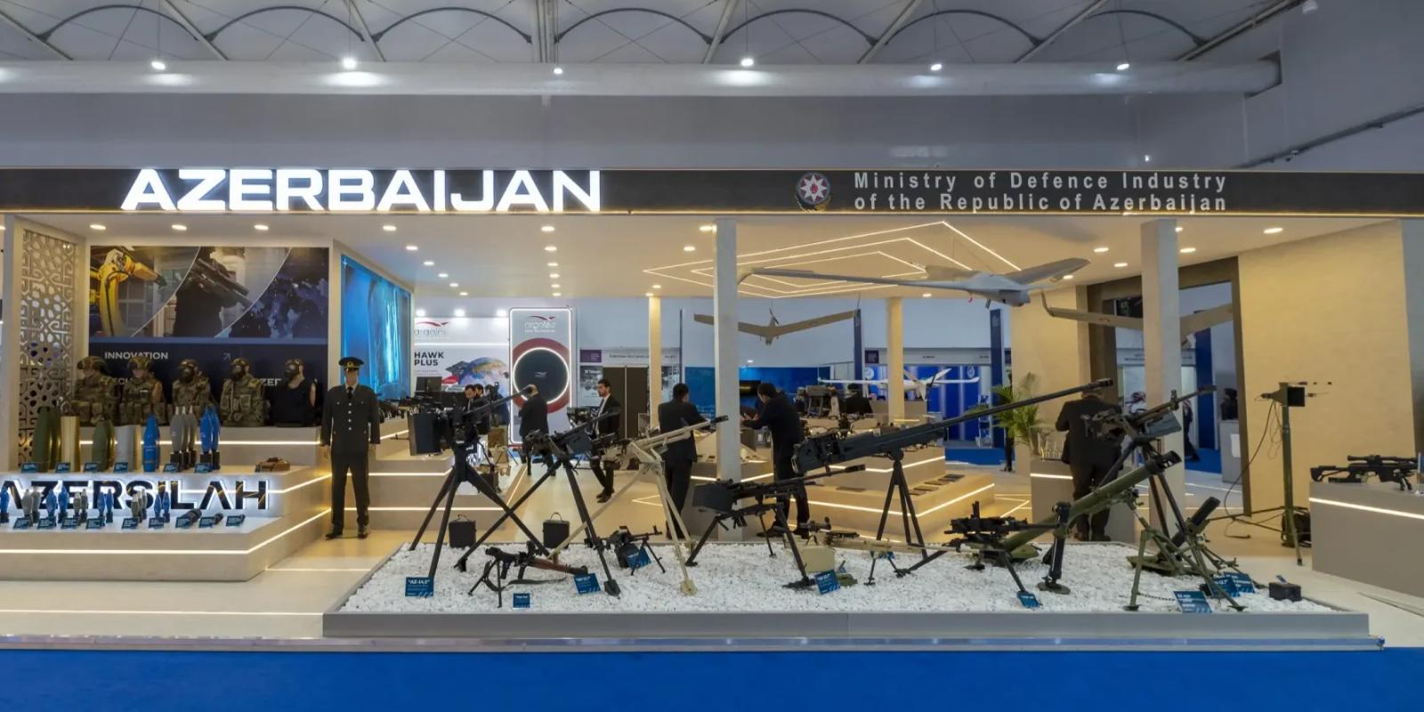 Front of the Azersilah national defence pavilion, with AZERBAIJAN and Ministry of Defence Industry of the Republic of Azerbaijan signage above uniformed mannequins and tripod-mounted weapons on white gravel