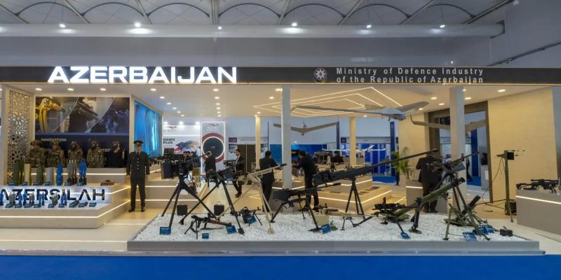 Front of the Azersilah national defence pavilion, with AZERBAIJAN and Ministry of Defence Industry of the Republic of Azerbaijan signage above uniformed mannequins and tripod-mounted weapons on white gravel