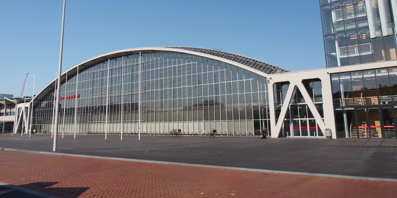 Arched glass facade of the Europahal at RAI Amsterdam on Europaplein, under a clear blue sky, with an empty paved forecourt