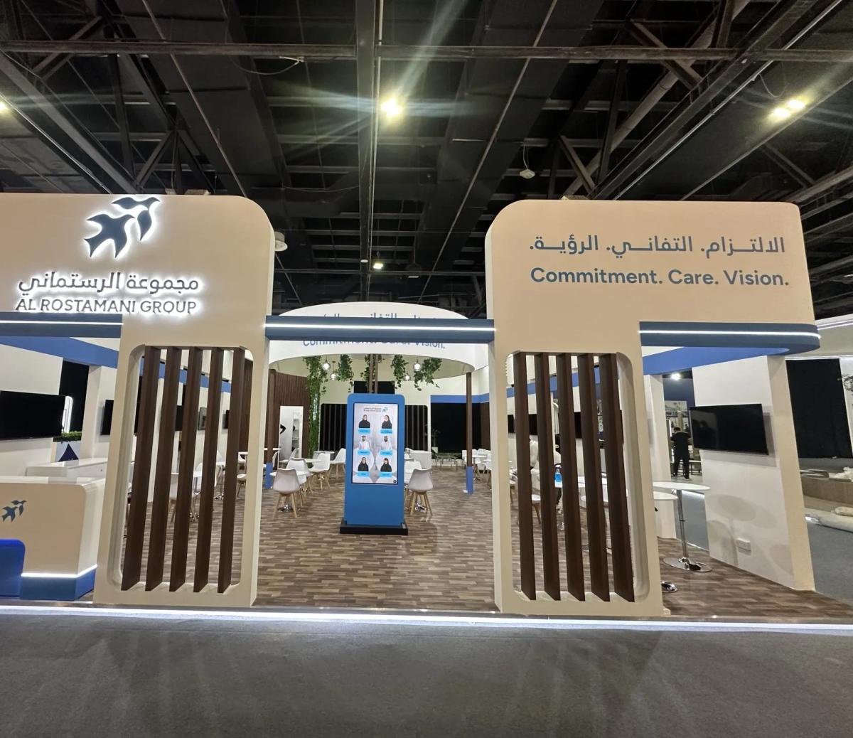 Al Rostamani Group exhibition stand with cream frames, dark wood-effect vertical slats, blue accent lighting and a digital display column at its centre
