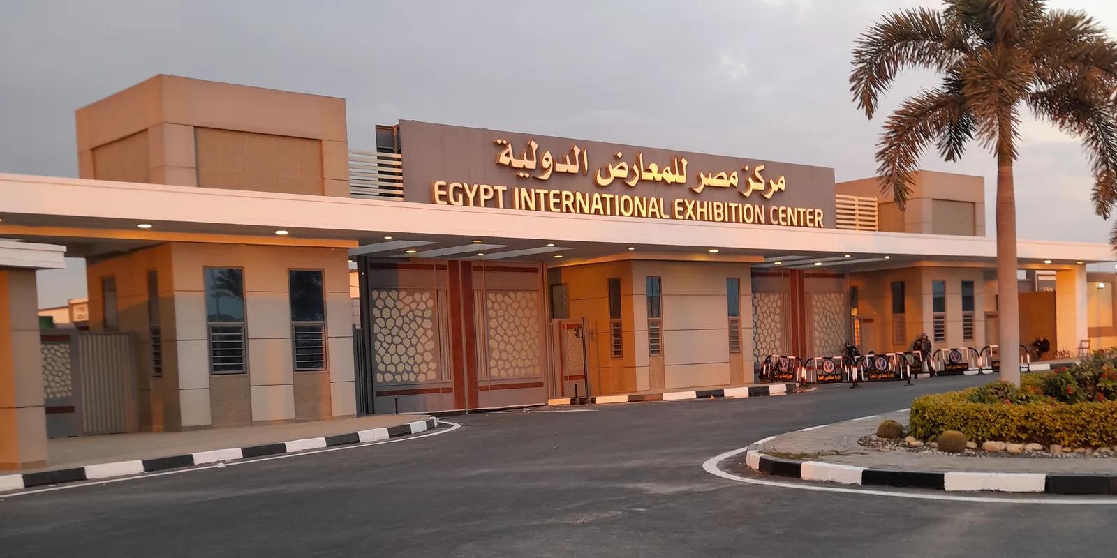 Entrance of the Egypt International Exhibition Centre with its Arabic and English name sign above the gates, palm trees and a curved driveway at dusk