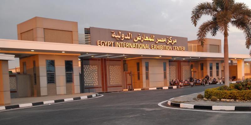 Entrance of the Egypt International Exhibition Centre with its Arabic and English name sign above the gates, palm trees and a curved driveway at dusk